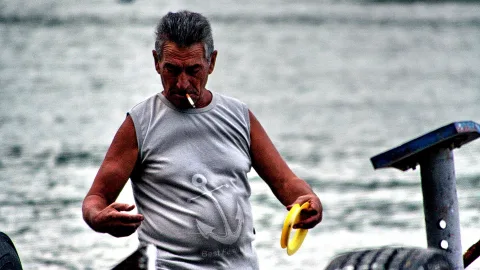 Älterer Mann mit grauen Haaren und ärmellosem grauem Shirt mit Anker-Motiv raucht eine Zigarette und hält einen gelben Ring in den Händen, im Hintergrund unscharfes Wasser.