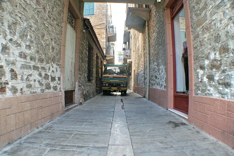 Schmale Gasse mit grob verputzten Steinmauern und rot gerahmten Türen, am Ende steht ein alter bunter LKW.