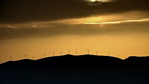 Silhouetten von Windrädern auf einem Hügelkamm vor goldgelbem Abendhimmel mit dunklen Wolken.