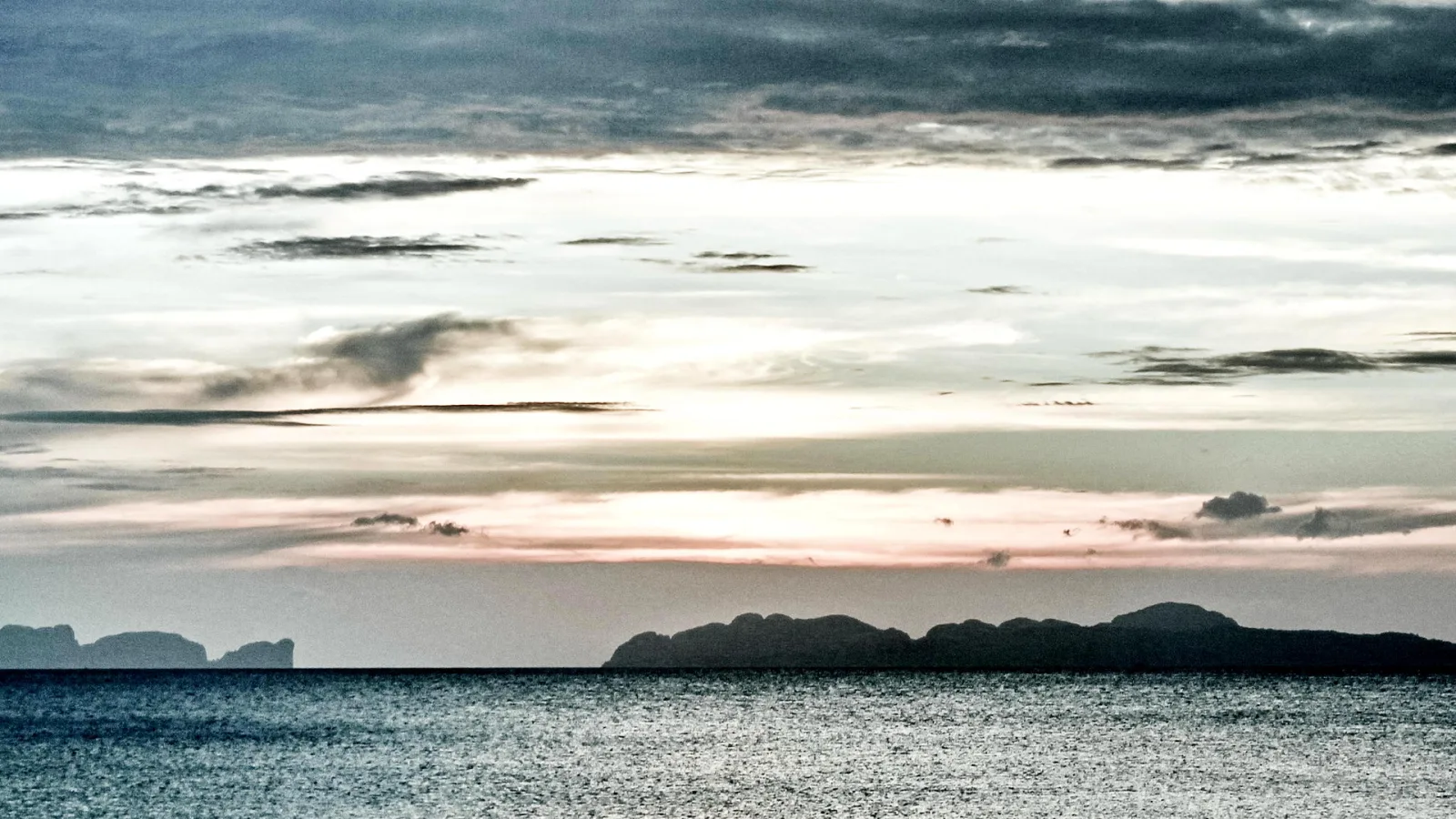 Weite Meeresfläche mit glitzernder Wasseroberfläche, darüber eine bewölkte Abendhimmel mit rosa Schimmer am Horizont, rechts und links am Horizont dunkle Felseninseln.