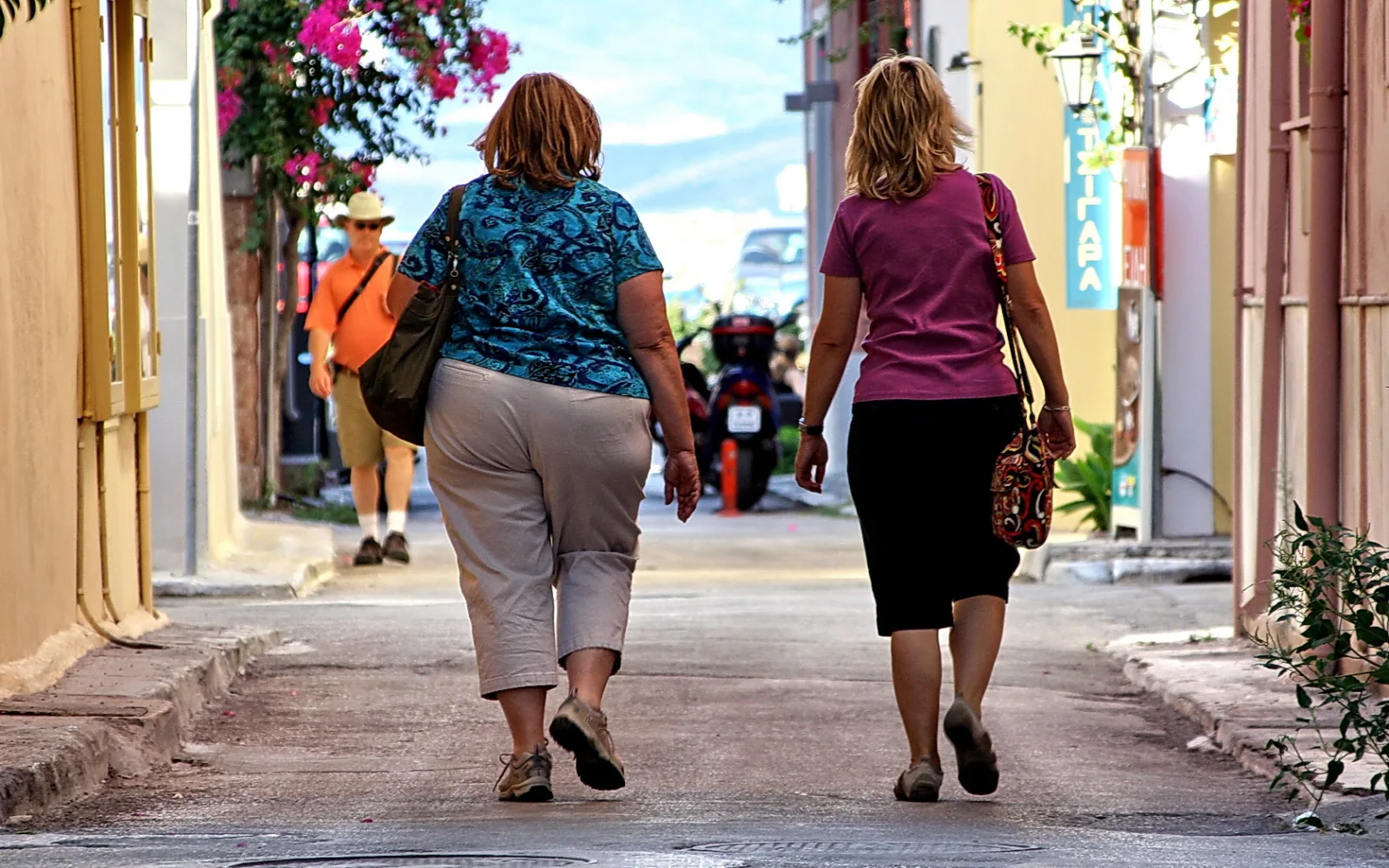 Zwei Frauen von hinten auf einer schmalen Gasse, links in blau gemusterter Bluse und Caprihose, rechts in pinkfarbenem Shirt und schwarzem Rock. Im Hintergrund geht ein Mann mit Hut und oranger Jacke.