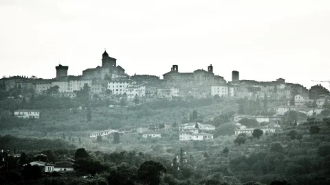 Ein auf einem Hügel gelegenes Dorf mit Kirchturm, Wehrturm und dicht stehenden Häusern, davor dunstige Baumlandschaft in gedämpften Grüntönen.
