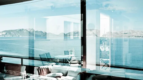 Blick durch eine große Glasfront auf das blaue Mittelmeer und eine bergige Küste, davor Terrassenmöbel, ein Tisch mit Blumenvase und ein weißes Sofa mit Kissen.
