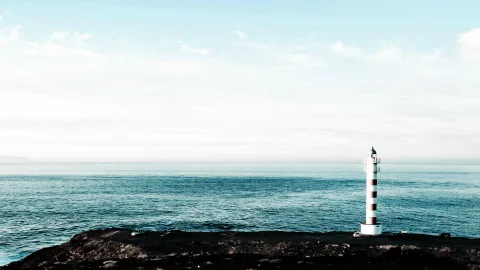 Weiß-rot gestreifter Leuchtturm auf einer dunklen Felsküste, davor ruhiges blaugrünes Meer unter hellem, diesigem Himmel.