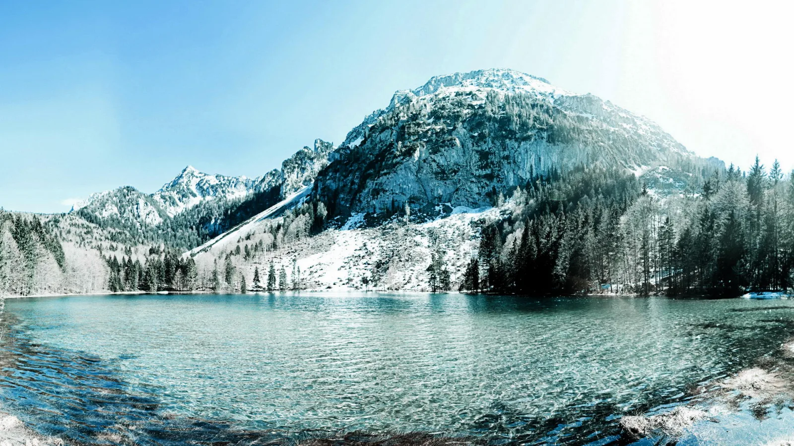 Ein türkisfarbener See vor einem verschneiten Bergmassiv, am Ufer dunkle Nadelbäume unter blauem Himmel.