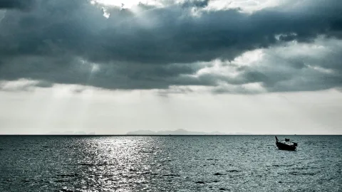 Ein einzelnes Langboot schwimmt auf ruhigem, silbrig glänzendem Meer, darüber türmen sich dunkle Wolken, durch die Sonnenstrahlen fallen. In der Ferne zeichnen sich flache Inseln am Horizont ab.