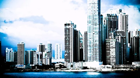 Skyline mit dicht stehenden Hochhäusern am Ufer, davor ein dunkles Wasser, darüber bewölkter blauer Himmel mit Dunst am Horizont.