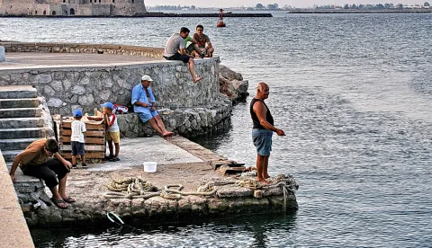 Männer und Kinder angeln und sitzen auf einer Hafenmauer am Wasser, im Hintergrund eine Festung.