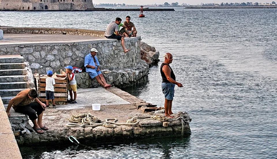 Männer und Kinder angeln und sitzen auf einer Hafenmauer am Wasser, im Hintergrund eine Festung.