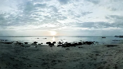 Breites Panorama eines flachen Sandstrands bei Sonnenuntergang, im Vordergrund dunkle Felsen und Fußspuren im Sand, rechts ein kleines Boot auf ruhigem Meer unter bewölktem Himmel.