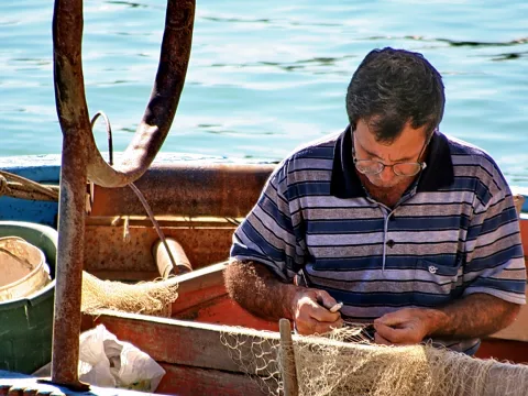 Ein Mann mit Brille und gestreiftem Polohemd sitzt im Fischerboot und flickt ein helles Netz, links steht ein rostiger Anker vor dem Wasser.