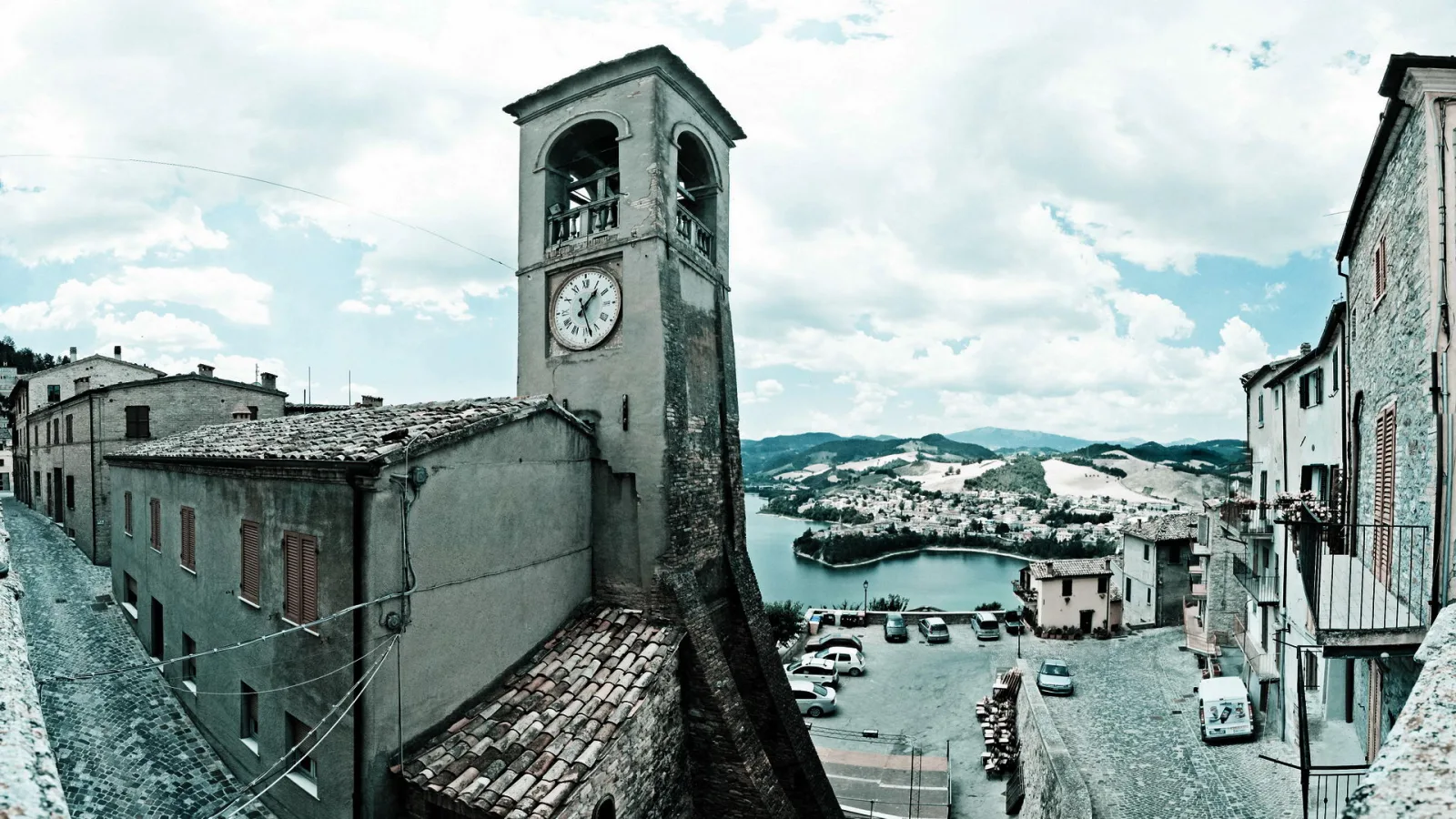 Ein Kirchturm mit Uhr ragt über alte Steinhäuser und eine Gasse, dahinter liegen ein See und bewaldete Hügel unter einem bewölkten Himmel.