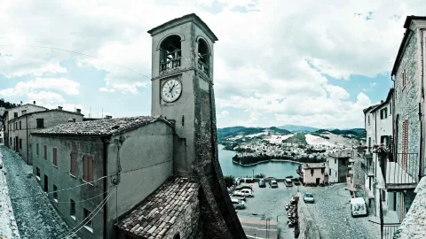 Ein Kirchturm mit Uhr ragt über alte Steinhäuser und eine Gasse, dahinter liegen ein See und bewaldete Hügel unter einem bewölkten Himmel.