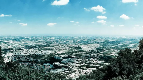 Weitblick von einem bewaldeten Hang auf eine hügelige Landschaft mit dicht bebauten Ortschaften, Feldern und blauem Himmel mit kleinen Wolken.