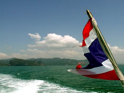 Rot-weiß-blau gestreifte thailändische Flagge weht am Heck eines Bootes, dahinter Meer und bewaldete Inseln unter blauem Himmel mit Wolken.