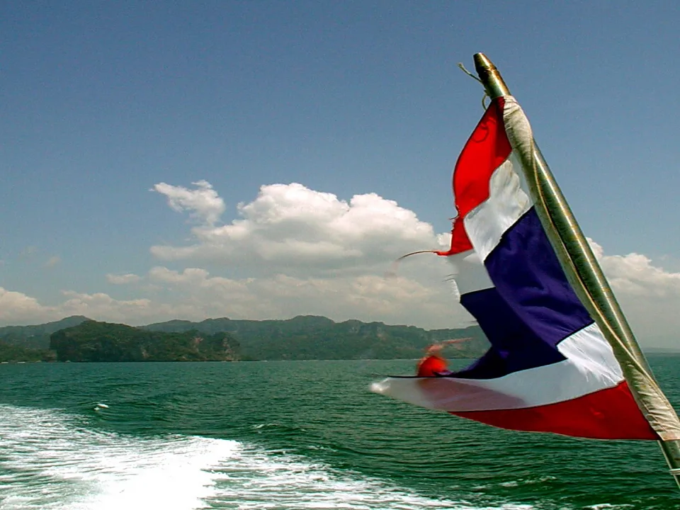 Rot-weiß-blau gestreifte thailändische Flagge weht am Heck eines Bootes, dahinter Meer und bewaldete Inseln unter blauem Himmel mit Wolken.
