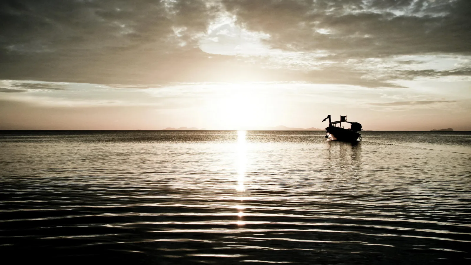 Ein dunkles Boot mit Aufbau fährt auf ruhigem Meer in Richtung der tief stehenden Sonne, deren Spiegelung eine helle Bahn auf dem Wasser bildet. Der Himmel ist bewölkt, am Horizont liegen flache Inseln.