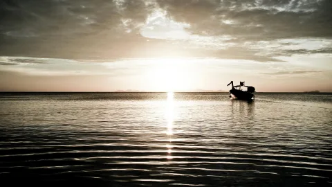 Ein dunkles Boot mit Aufbau fährt auf ruhigem Meer in Richtung der tief stehenden Sonne, deren Spiegelung eine helle Bahn auf dem Wasser bildet. Der Himmel ist bewölkt, am Horizont liegen flache Inseln.