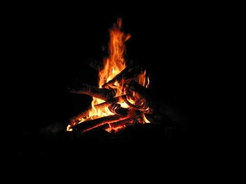 Brennendes Lagerfeuer mit aufgeschichteten Holzscheiten vor schwarzem Hintergrund, die Flammen leuchten orange.
