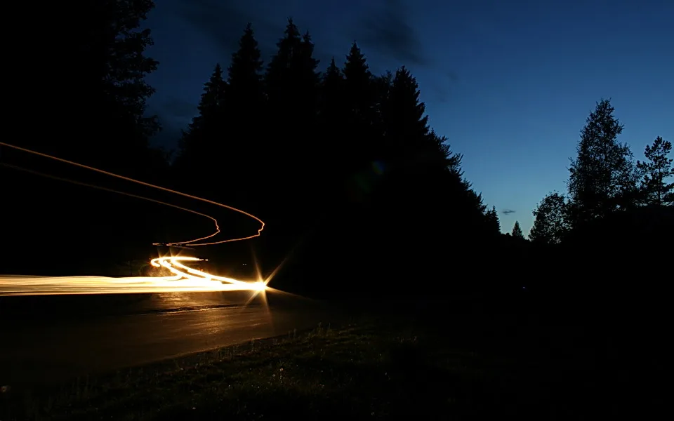 Langzeitbelichtung einer nächtlichen Straßenkurve, Scheinwerferlicht zieht als helle Spur nach links, dunkle Nadelbäume säumen die Straße unter blauem Abendhimmel.
