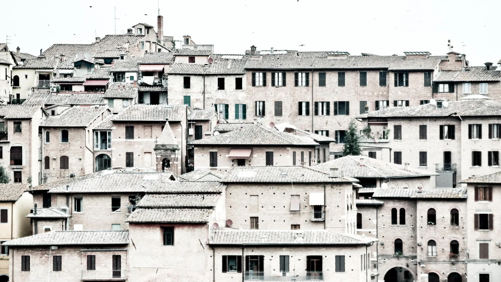 Dicht gedrängte Backsteinhäuser mit Ziegeldächern und Fensterläden in der Altstadt von Siena unter hellem, bedecktem Himmel.
