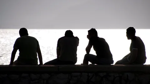 Vier Männer sitzen als dunkle Silhouetten nebeneinander auf einer Steinmauer, vor einer hell glitzernden Wasserfläche und grauem Himmel.
