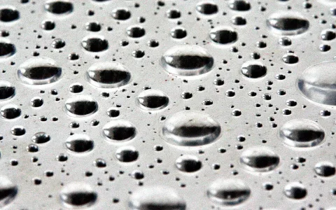 Viele Wassertropfen unterschiedlicher Größe auf einer glatten, grauen Oberfläche mit hellen Spiegelungen.
