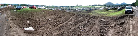 Panoramaaufnahme eines matschigen, von Reifenspuren durchzogenen Geländes, auf dem zahlreiche Autos parken; im Hintergrund bewaldete Hügel unter bewölktem Himmel.