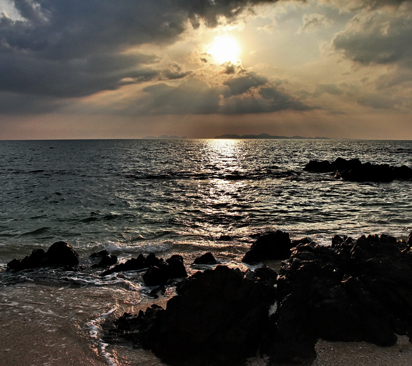 Dunkle Felsen am Strand im Vordergrund, dahinter ein Meer mit Sonnenreflexion und die tief hängende Sonne hinter Wolken am Horizont.