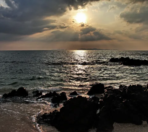 Dunkle Felsen am Strand im Vordergrund, dahinter ein Meer mit Sonnenreflexion und die tief hängende Sonne hinter Wolken am Horizont.