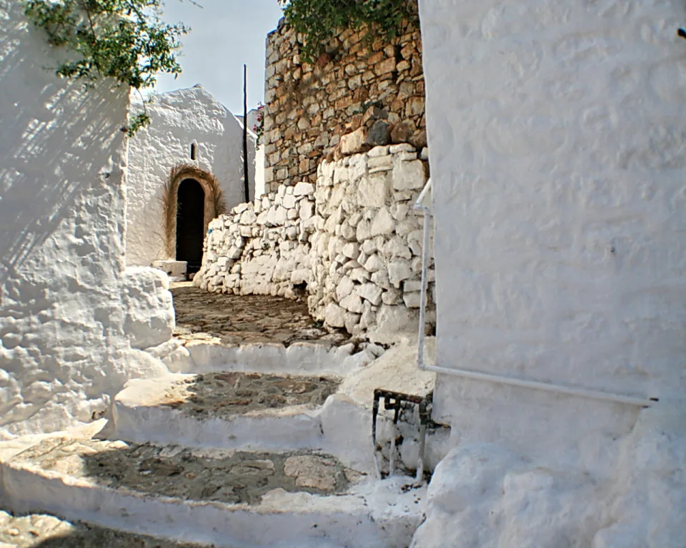 Schmale Gasse mit weiß getünchten Mauern, einer Steinmauer links im Hintergrund und Stufen aus Naturstein, die zu einem gewölbten Eingang führen.