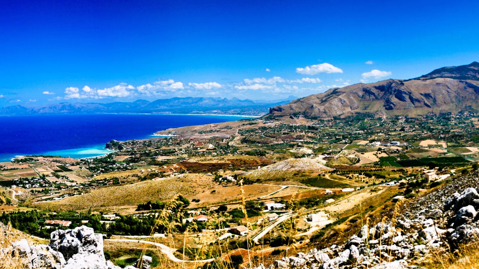 Blick über eine Küstenbucht mit tiefblauem Meer, Bergen und verstreuten Häusern in Sizilien.