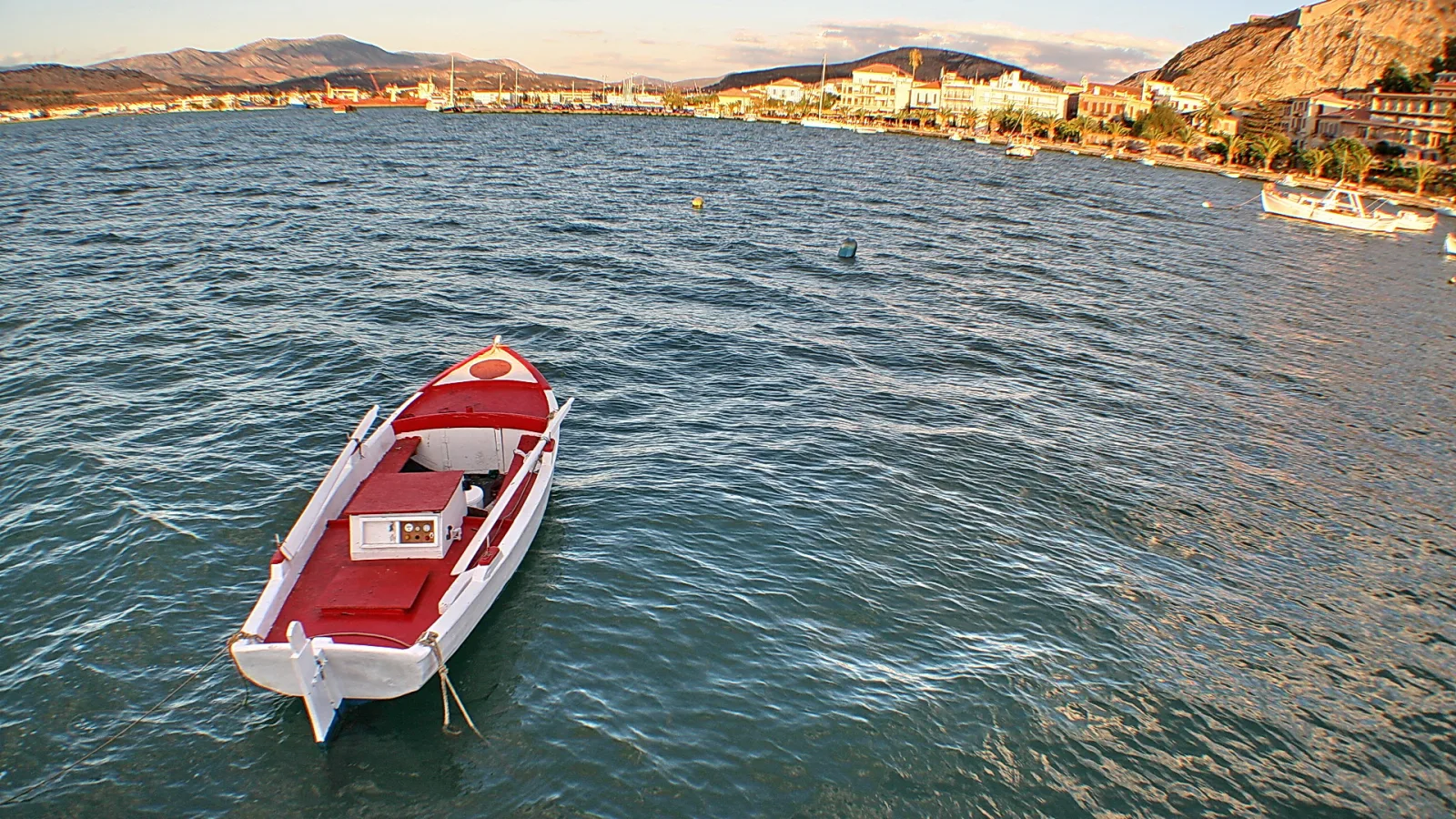 Ein rot-weißes Ruderboot liegt auf ruhigem, blaugrünem Wasser vor einer Hafenstadt mit Hügeln im Hintergrund.