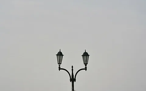 Laternenmast mit zwei symmetrisch angeordneten schwarzen Laternen vor einem hellgrauen, wolkenverhangenen Himmel