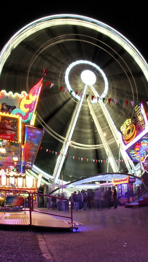 Ein beleuchtetes Riesenrad vor dunklem Himmel, im Vordergrund Jahrmarktsbuden mit bunten Lichtern und eine Gruppe Menschen, die Bewegung ist durch die Langzeitbelichtung verwischt.