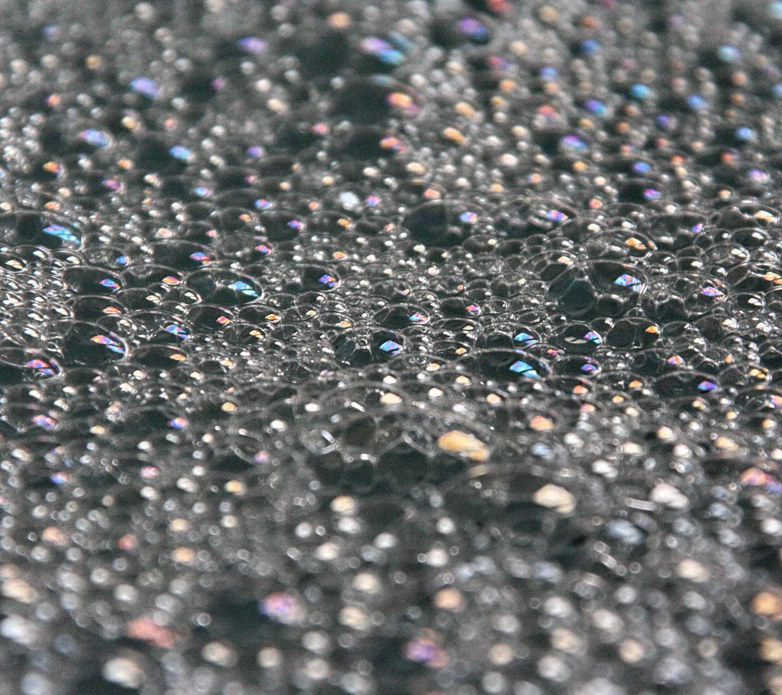 Nahaufnahme einer dunklen Oberfläche, bedeckt mit vielen kleinen Wasserbläschen, die im Licht bunt schillern.