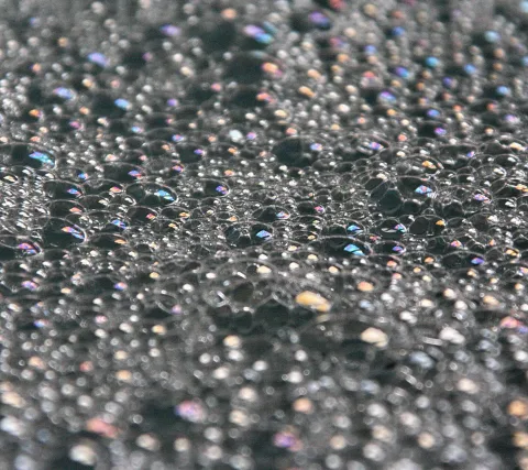 Nahaufnahme einer dunklen Oberfläche, bedeckt mit vielen kleinen Wasserbläschen, die im Licht bunt schillern.