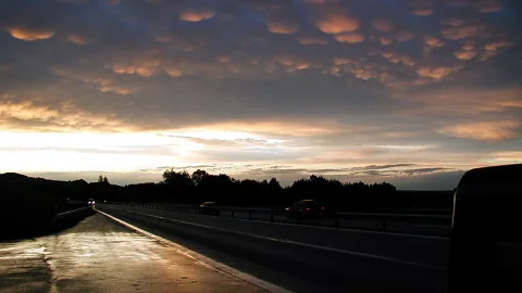 Autobahn bei Sonnenuntergang unter einem Himmel mit gewellten Wolkenbänken, die von unten orange angestrahlt werden. Rechts im Bild ragt ein dunkles Fahrzeug ins Bild, links führt der nasse Asphalt in die Ferne.