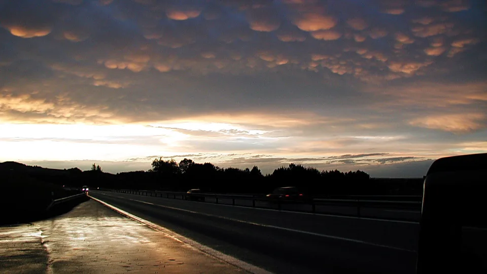 Autobahn bei Sonnenuntergang unter einem Himmel mit gewellten Wolkenbänken, die von unten orange angestrahlt werden. Rechts im Bild ragt ein dunkles Fahrzeug ins Bild, links führt der nasse Asphalt in die Ferne.