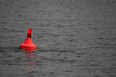Eine leuchtend rote Boje ragt links unten aus einer dunkelgrauen, leicht gekräuselten Wasserfläche.