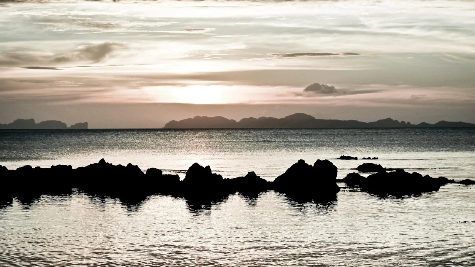 Dunkle Felsen zeichnen sich vor einem Meer ab, im Hintergrund liegen flache Inseln unter einem hellen Abendhimmel mit Sonne am Horizont.