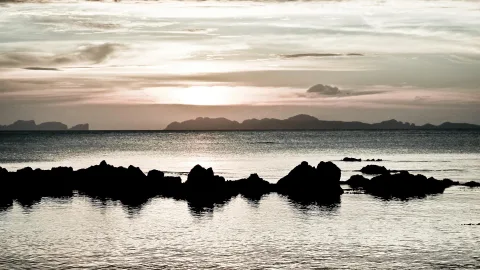 Dunkle Felsen zeichnen sich vor einem Meer ab, im Hintergrund liegen flache Inseln unter einem hellen Abendhimmel mit Sonne am Horizont.