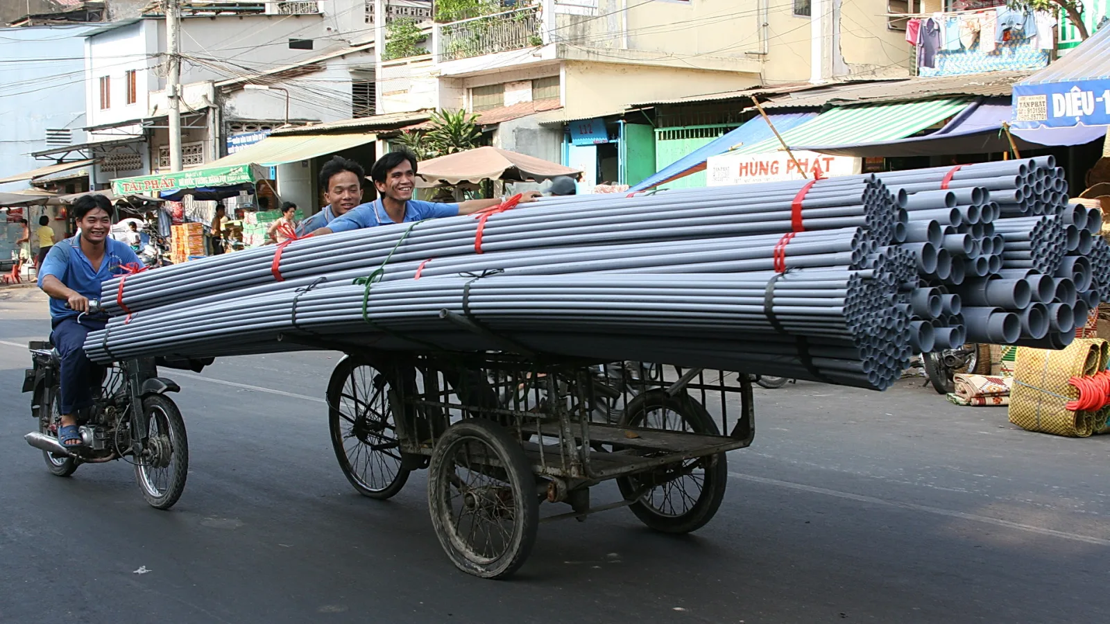 Drei Männer transportieren auf einem Motorrad und einem Handkarren einen hohen Stapel grauer Rohre über eine Straße in Ho-Chi-Minh-Stadt.