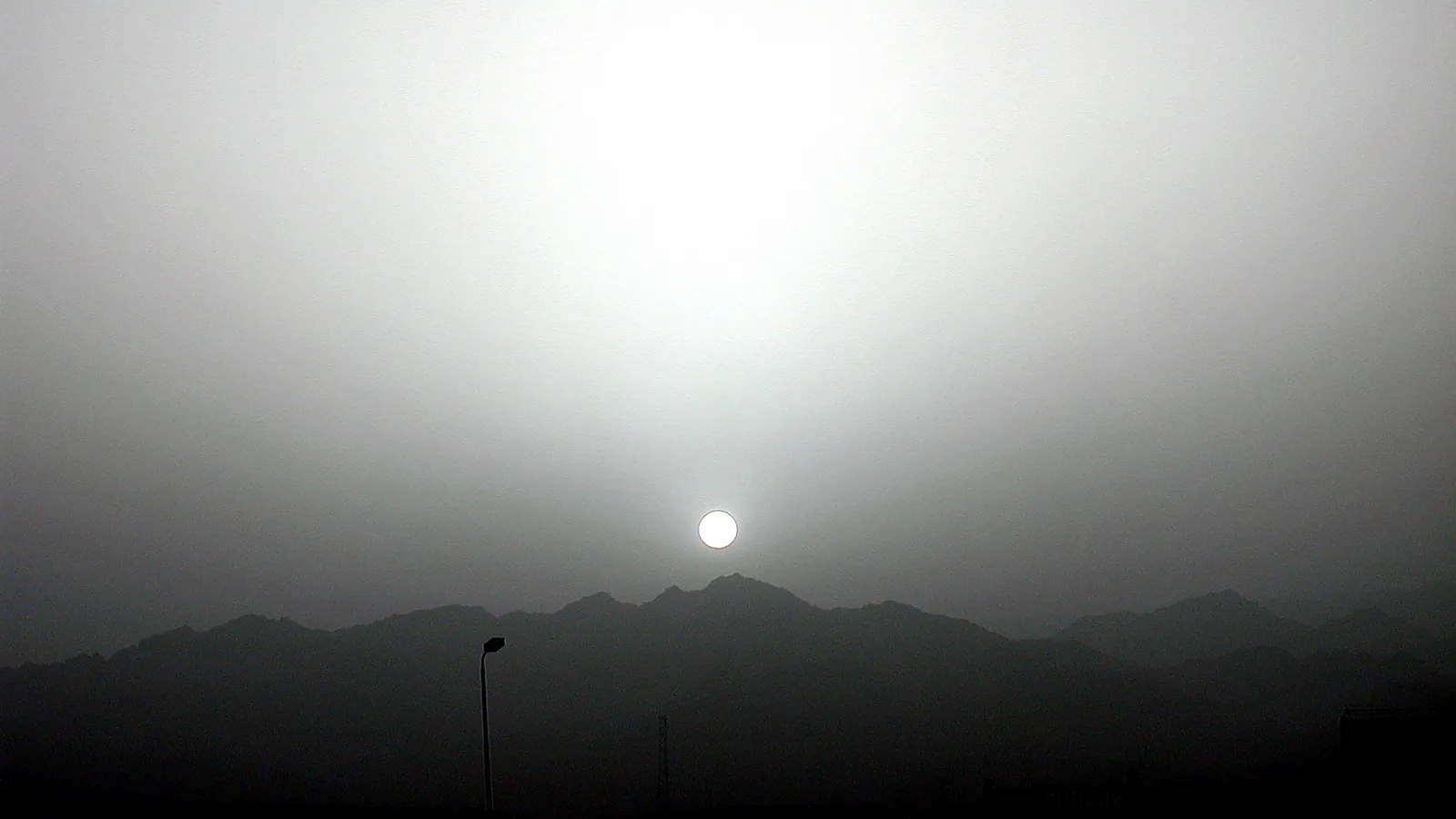 Eine blasse Sonne steht im Dunst knapp über einer dunklen Bergkette. Links ragt eine Straßenlaterne ins Bild, der Himmel ist grau und hell.