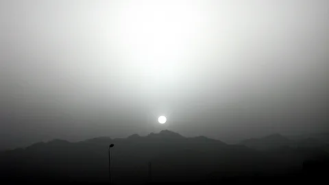 Eine blasse Sonne steht im Dunst knapp über einer dunklen Bergkette. Links ragt eine Straßenlaterne ins Bild, der Himmel ist grau und hell.