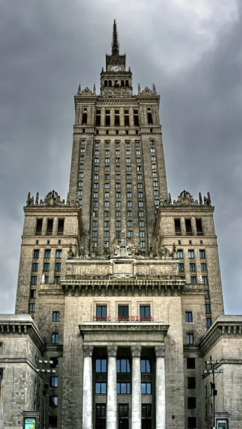 Symmetrische Fassade des Palastes der Kultur und Wissenschaft in Warschau, aufragend vor grauem, bewölktem Himmel, mit Säulenportal und Turm mit Uhr.