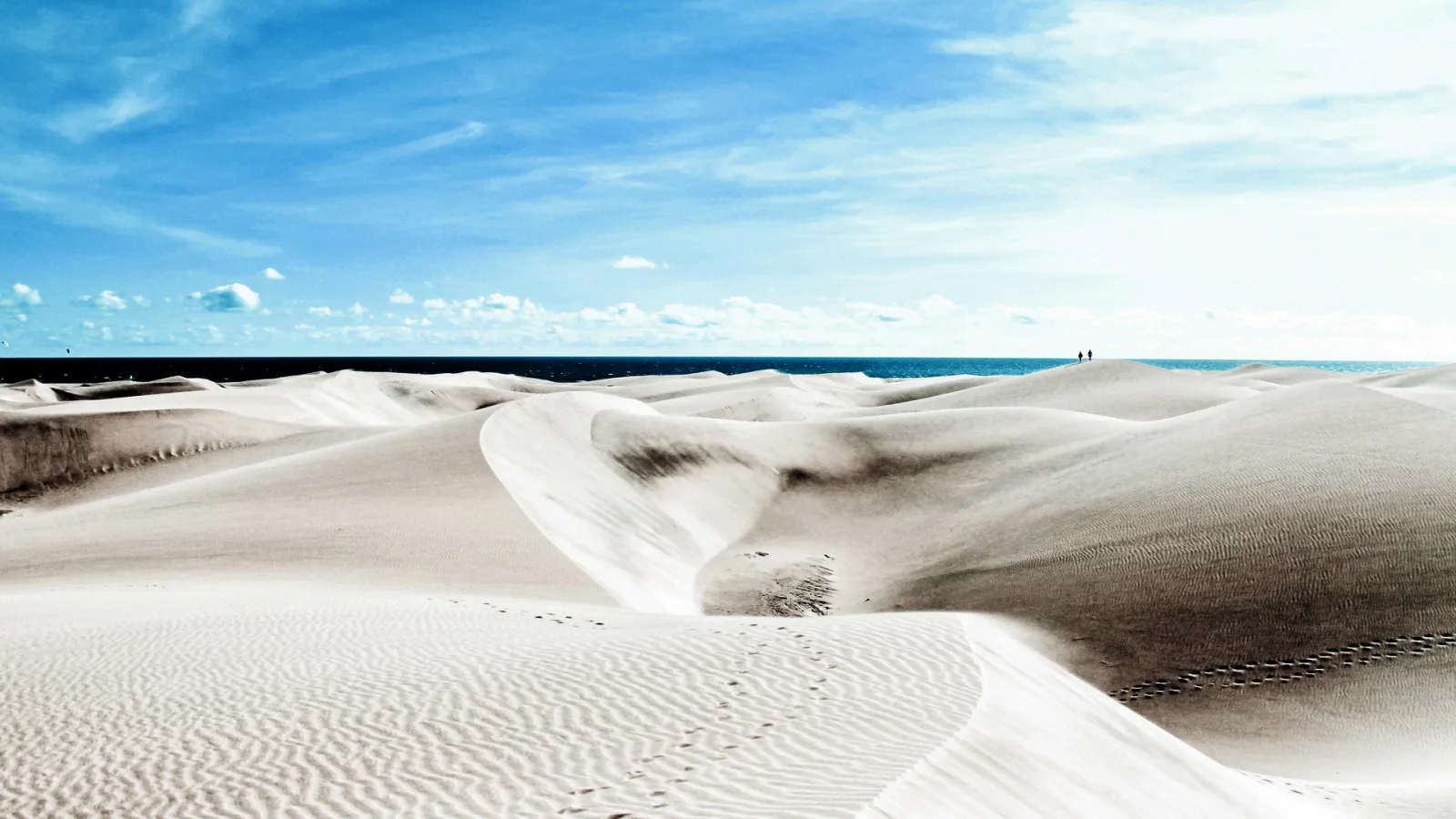 Helle Sanddünen unter blauem Himmel, dahinter das Meer am Horizont, im Vordergrund Fußspuren im Sand.