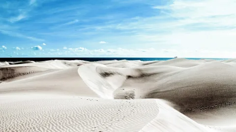 Helle Sanddünen unter blauem Himmel, dahinter das Meer am Horizont, im Vordergrund Fußspuren im Sand.