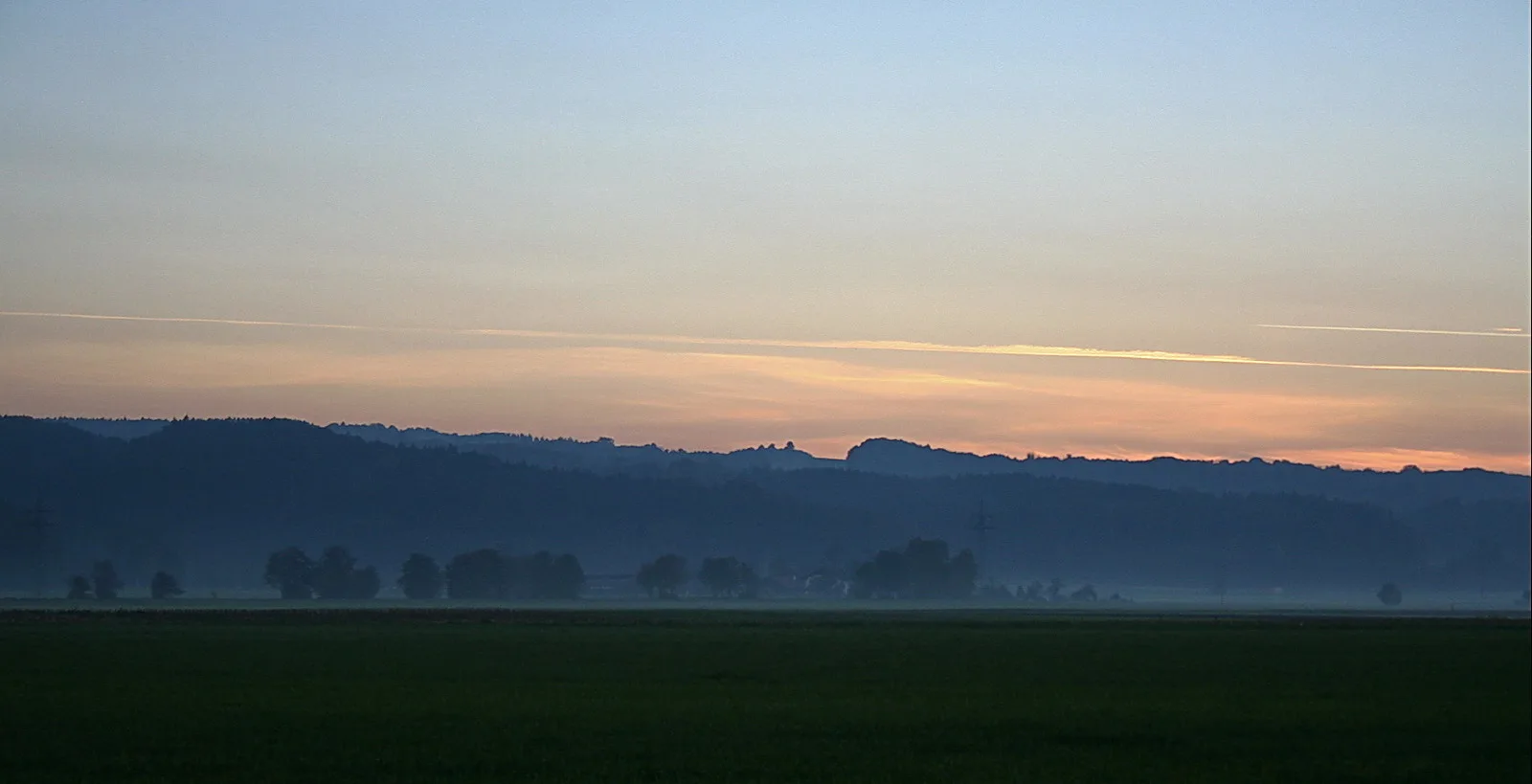 Diesige Landschaft am Abend mit bewaldeten Hügeln, Baumgruppen im Nebel und einem dunklen Feld im Vordergrund, darüber ein blassblau bis orange verlaufender Himmel mit Kondensstreifen.