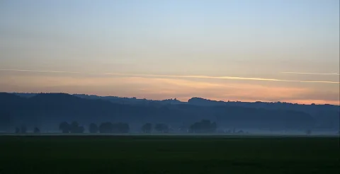 Diesige Landschaft am Abend mit bewaldeten Hügeln, Baumgruppen im Nebel und einem dunklen Feld im Vordergrund, darüber ein blassblau bis orange verlaufender Himmel mit Kondensstreifen.